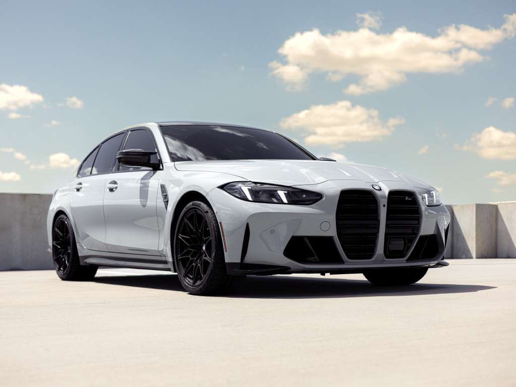 2026 BMW M3 Competition in light gray with black wheels against a blue sky backdrop.