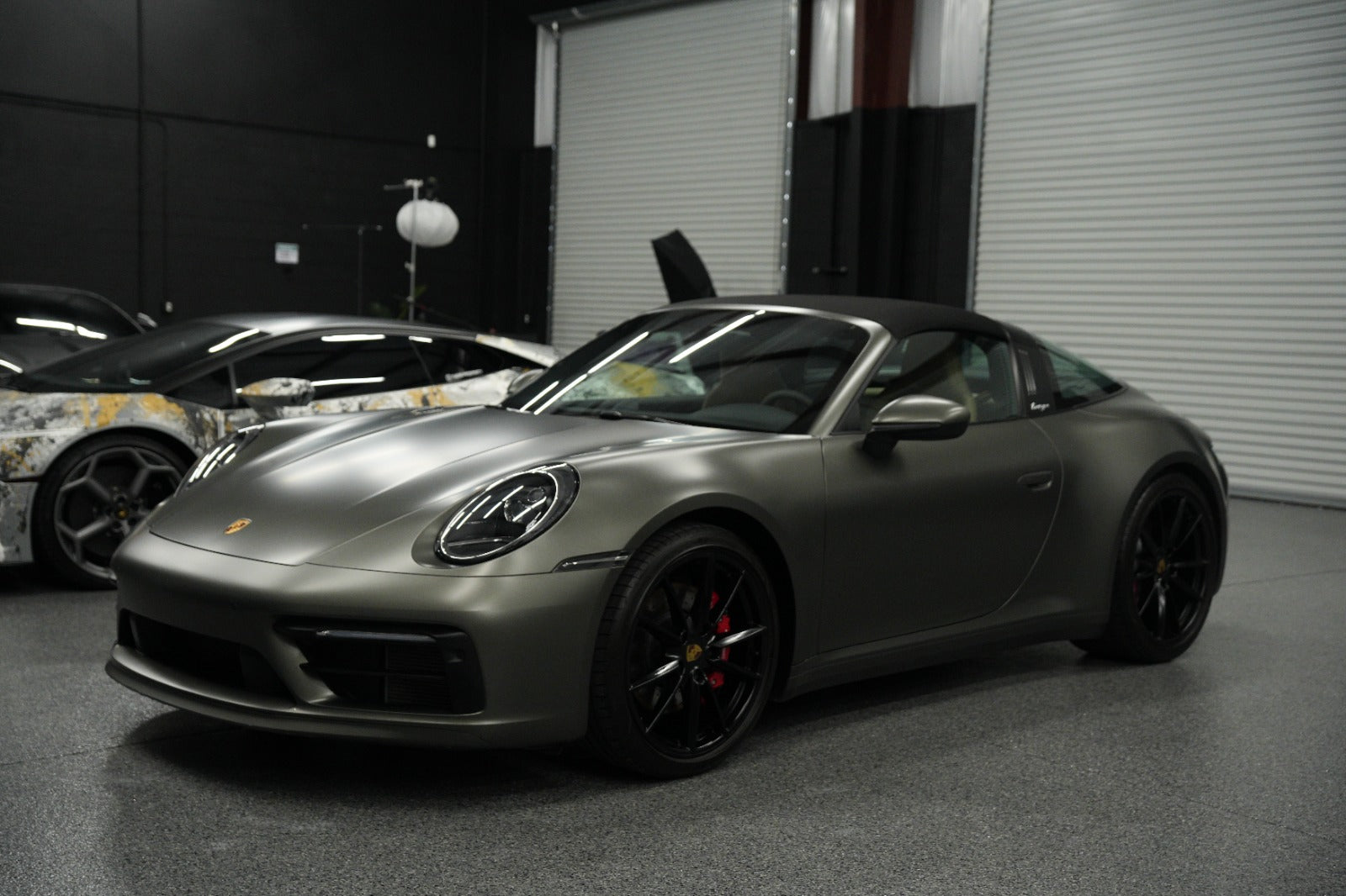 Matte gray Porsche 911 Carrera convertible sports car with black alloy wheels, red brake calipers, and sleek aerodynamic design parked in a garage showroom.