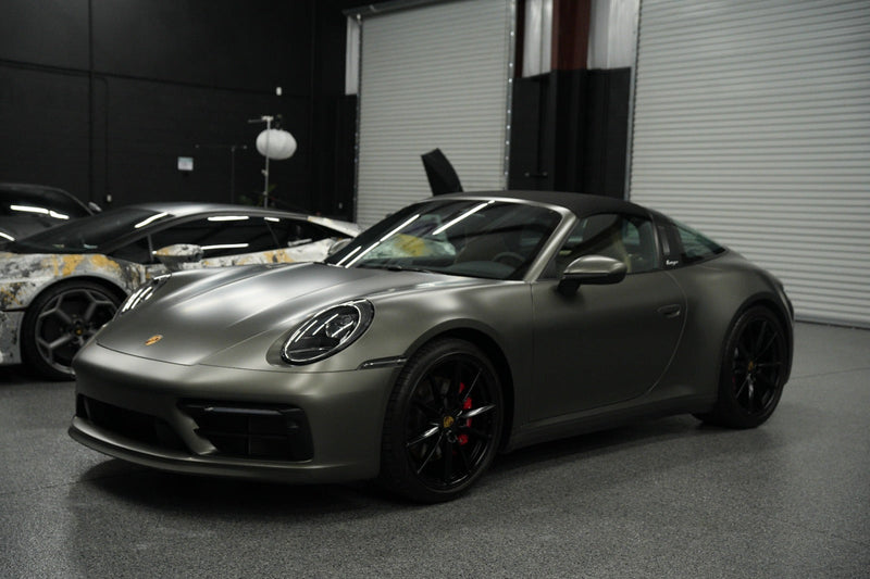 Matte gray Porsche 911 Carrera convertible sports car with black alloy wheels, red brake calipers, and sleek aerodynamic design parked in a garage showroom.