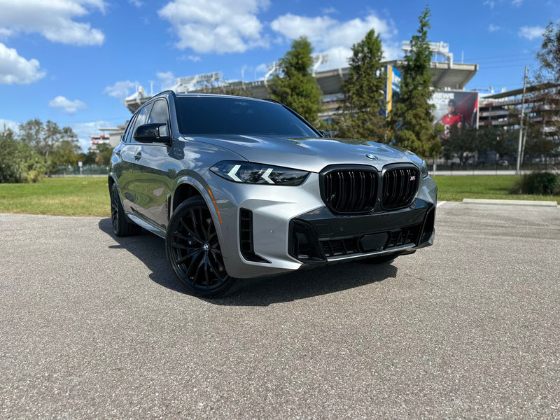 2023 BMW X5 M Sport utility vehicle in sleek metallic gray with black alloy wheels, parked outdoors under a sunny blue sky.