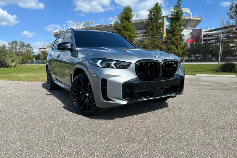 Silver BMW X5 M50i luxury SUV with black rims parked outdoors under clear blue sky, premium performance vehicle.