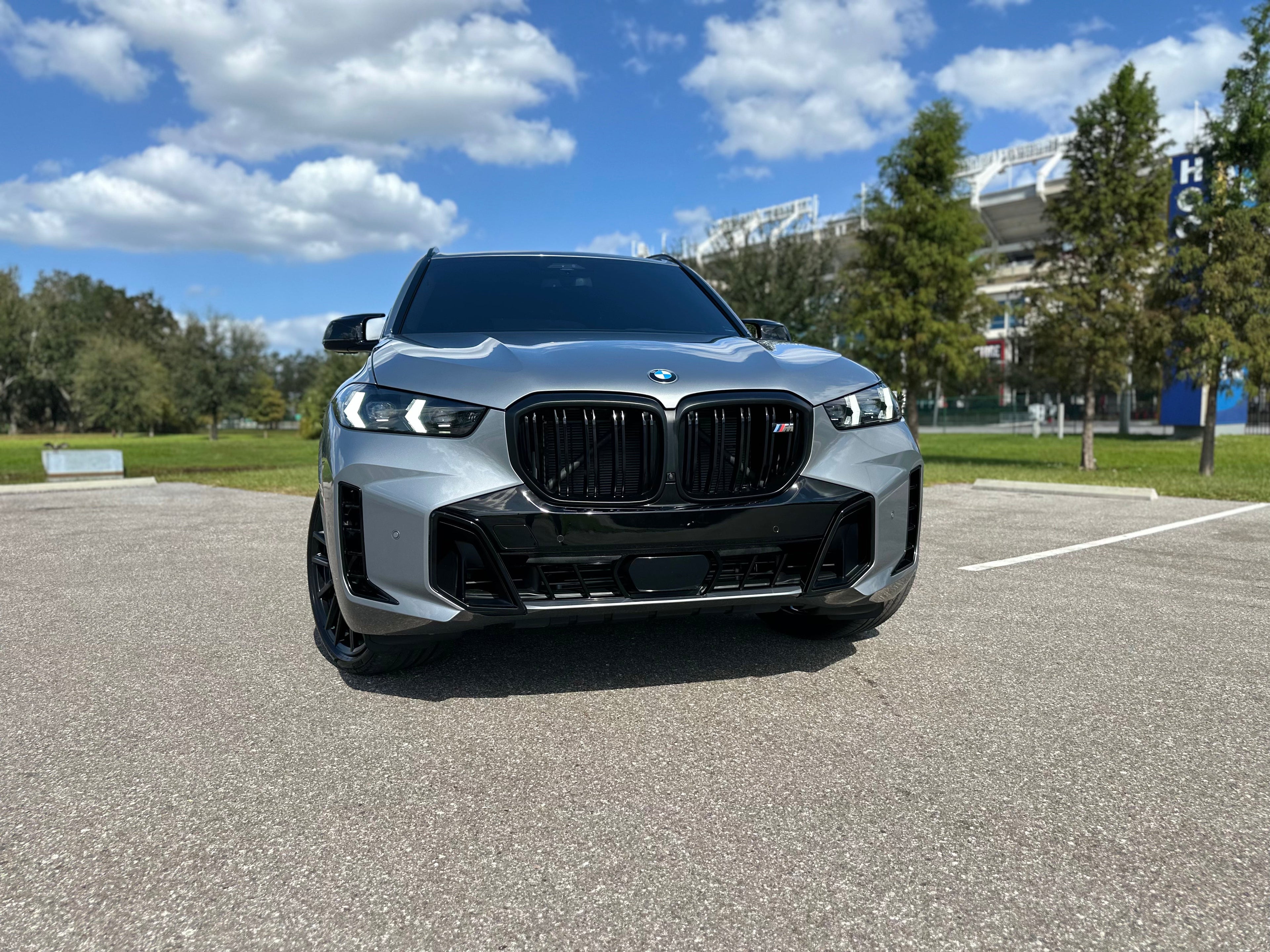 Front view of silver BMW X6 M50i with black kidney grille and LED headlights parked in outdoor lot under blue sky, luxury SUV exterior design.