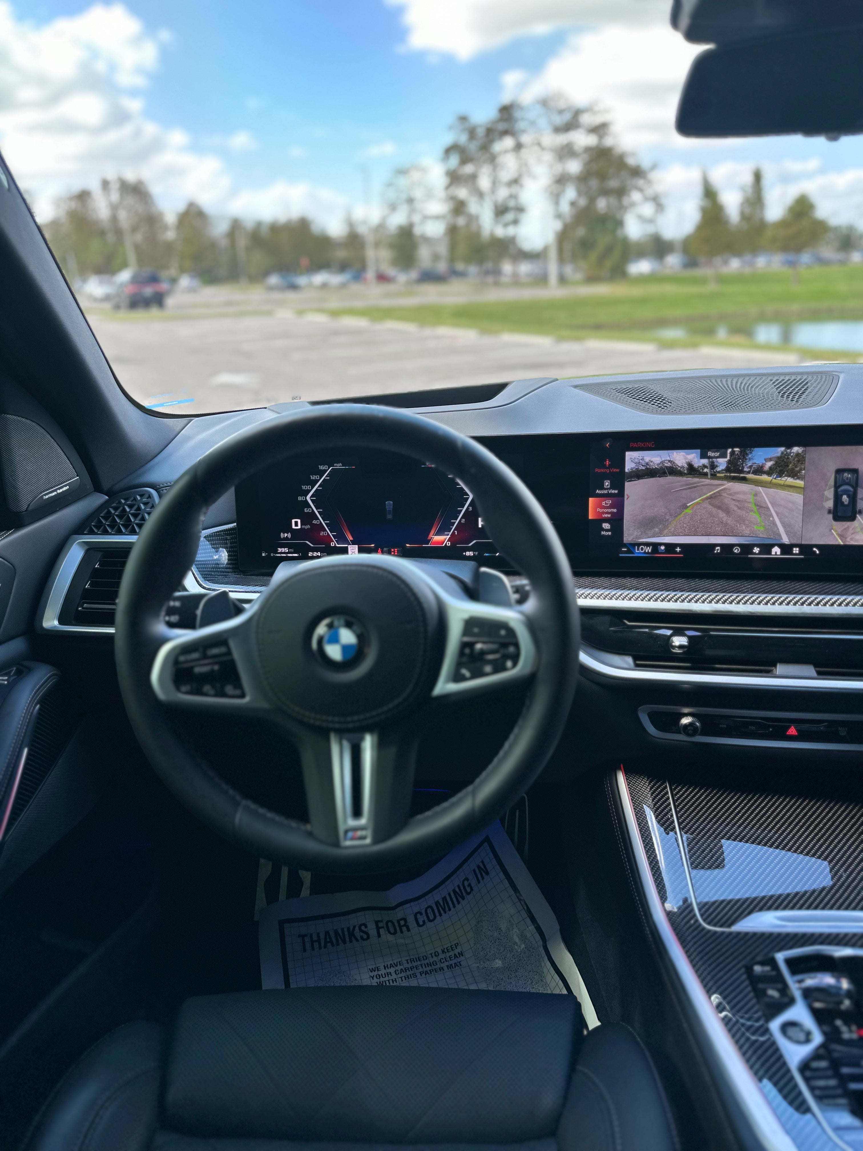 Interior view of BMW luxury car cockpit featuring leather steering wheel with BMW logo, digital dashboard display, and advanced touchscreen infotainment system with rearview camera, showcasing sleek black leather seats and carbon fiber trim accents.
