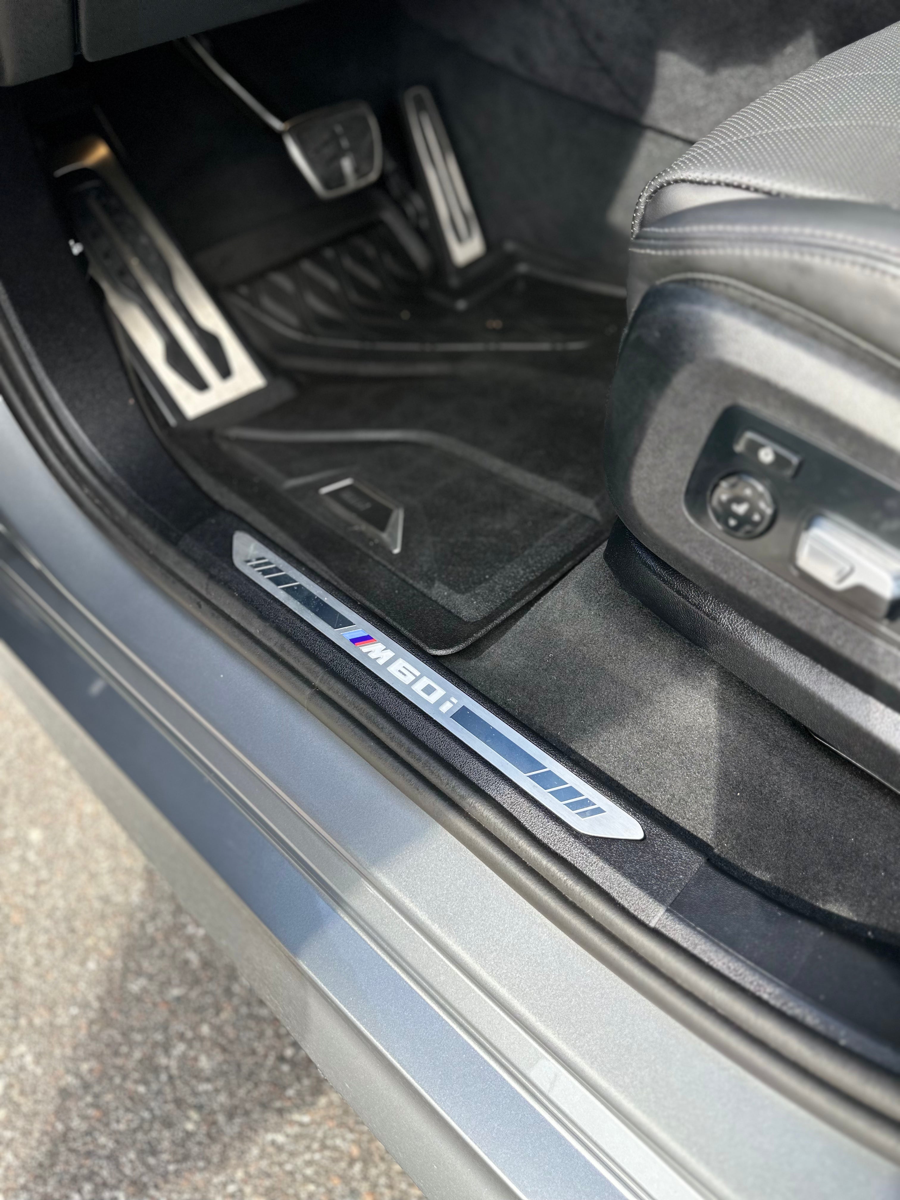 Close-up of BMW M60i car door sill plate and black floor mats, showcasing luxury interior details and sport pedal covers.