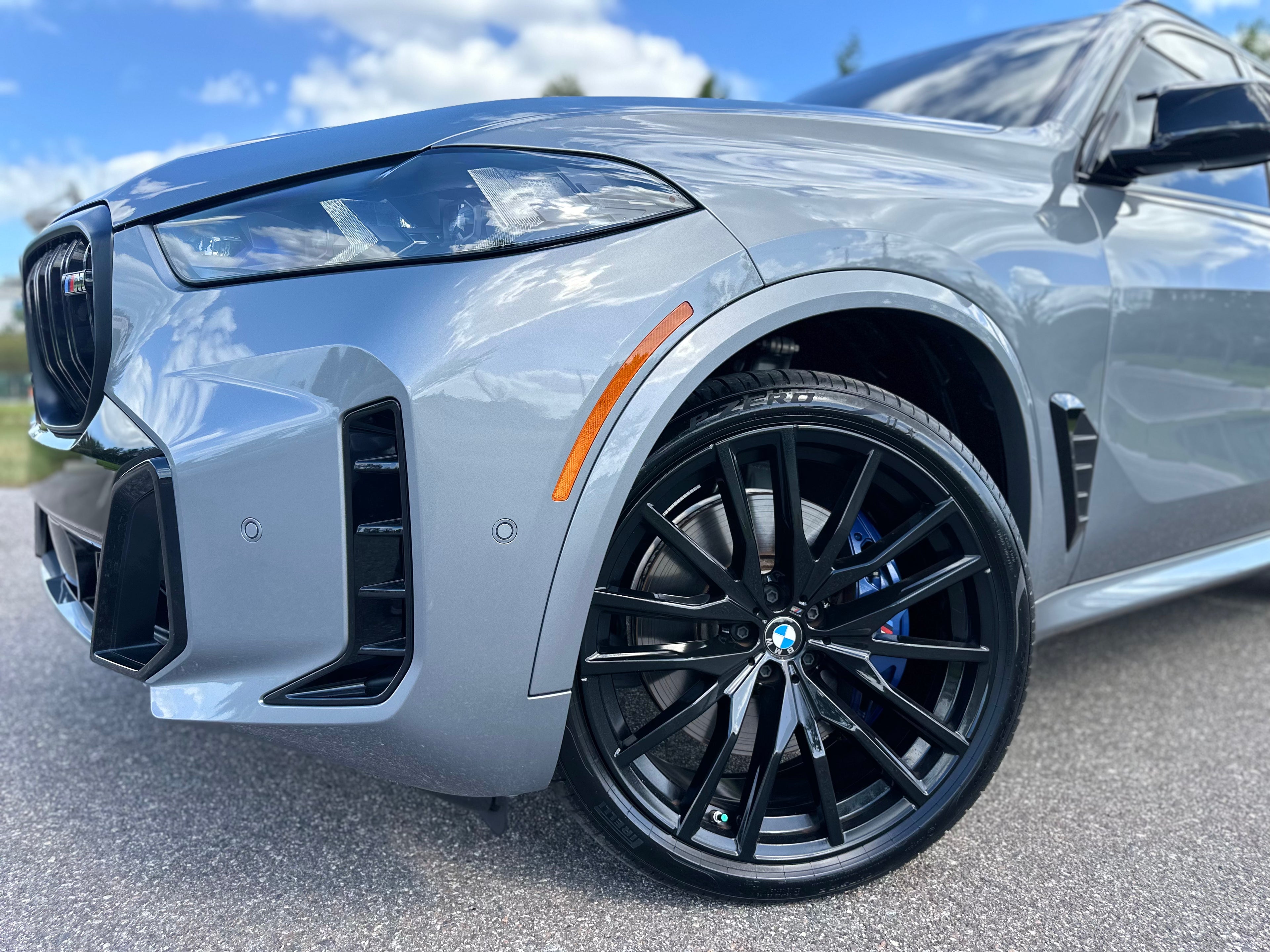 Close-up of silver BMW X5 M with black alloy wheels, Pirelli P Zero tires, and sporty front bumper design under clear blue sky.