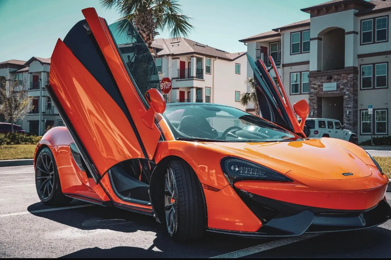 Orange luxury sports car with scissor doors open, parked in urban residential area, high-performance convertible with sleek aerodynamic design and black alloy wheels.