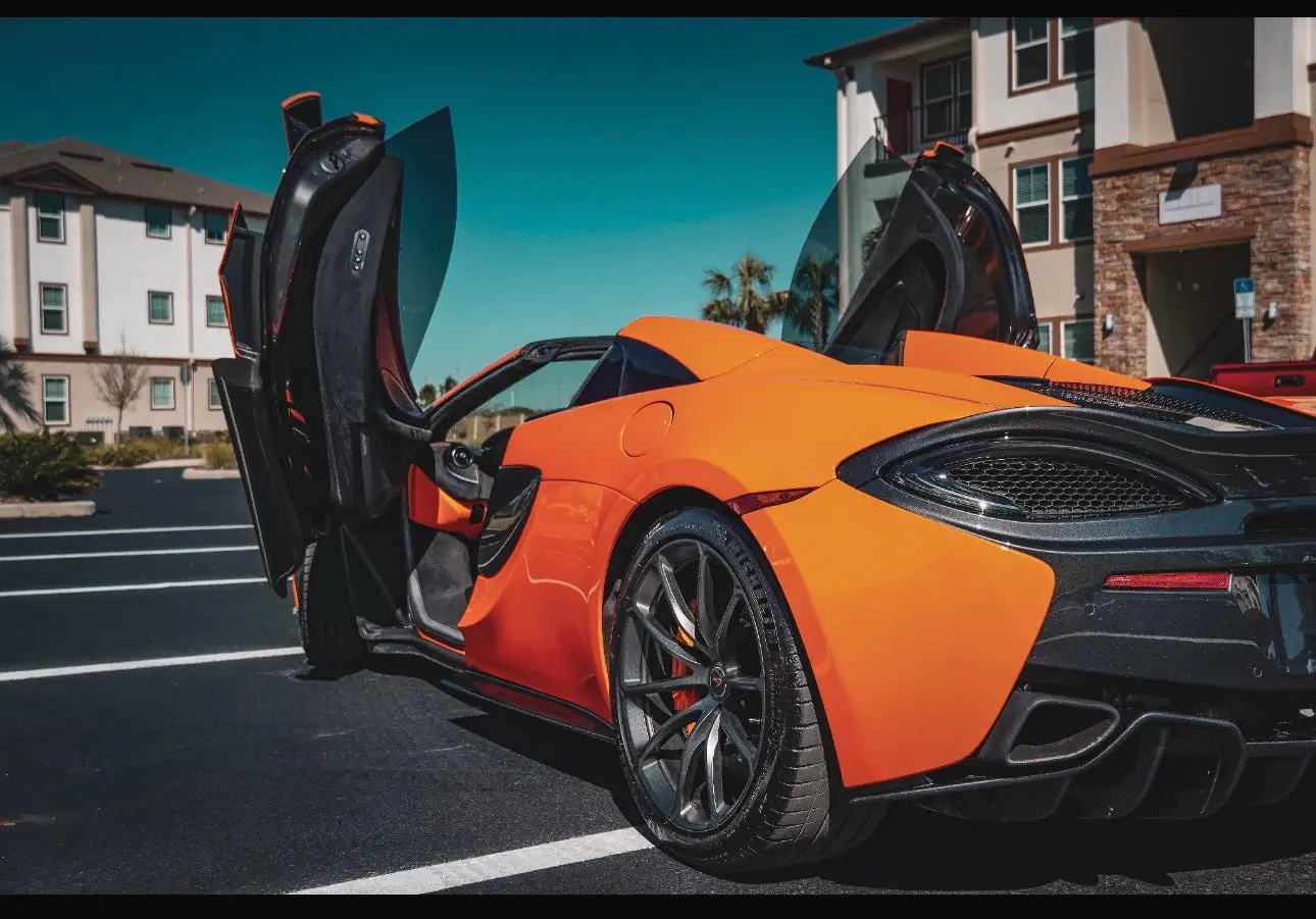 Orange luxury sports car with black alloy wheels and butterfly doors open, parked in a modern residential area, high-performance supercar design.