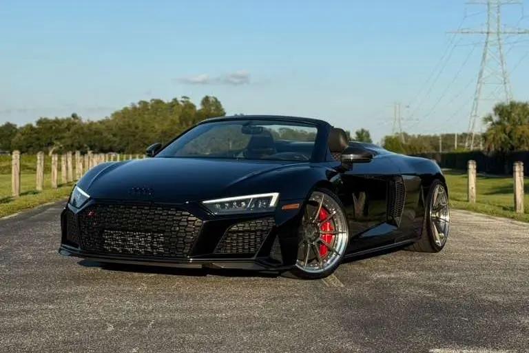 Black Audi R8 Spyder convertible sports car with red brake calipers, sleek aerodynamic design, and alloy wheels parked on open road.
