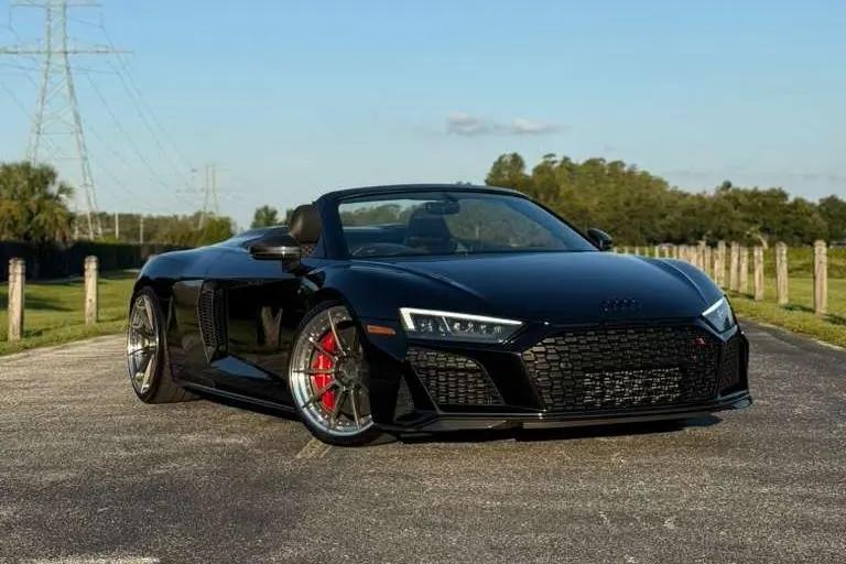 Black Audi R8 Spyder convertible sports car with red brake calipers, sleek aerodynamic design, and alloy wheels parked on open road.