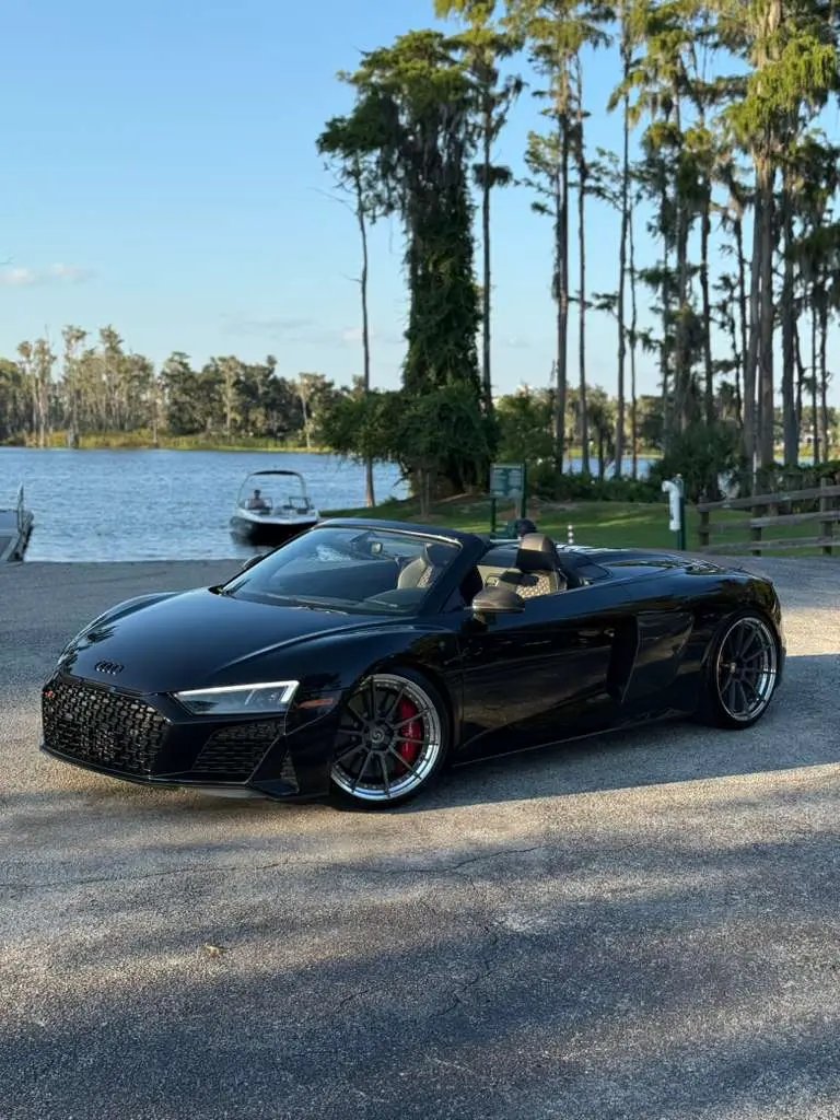 Black Audi R8 Spyder luxury convertible sports car parked near lake with trees, featuring sleek aerodynamic design, premium alloy wheels, and high-performance brakes.