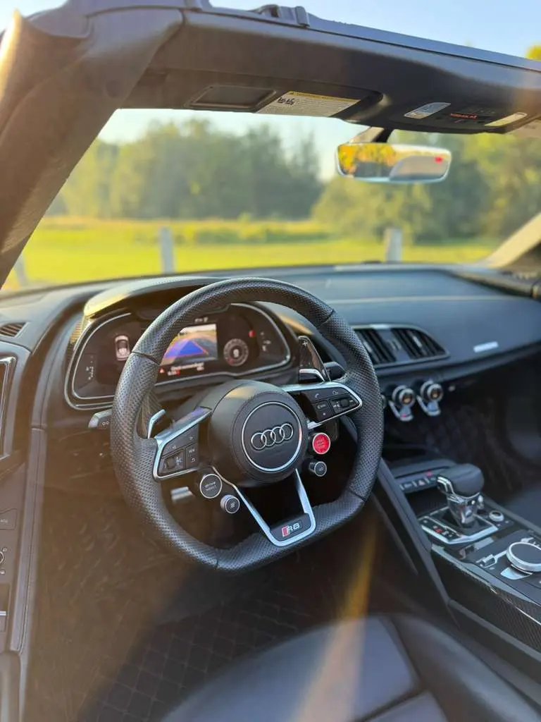 Interior view of a luxury Audi R8 sports car featuring a black perforated leather steering wheel with control buttons, modern digital dashboard, and sleek center console with gear shift and climate control, showcasing premium car interior design.