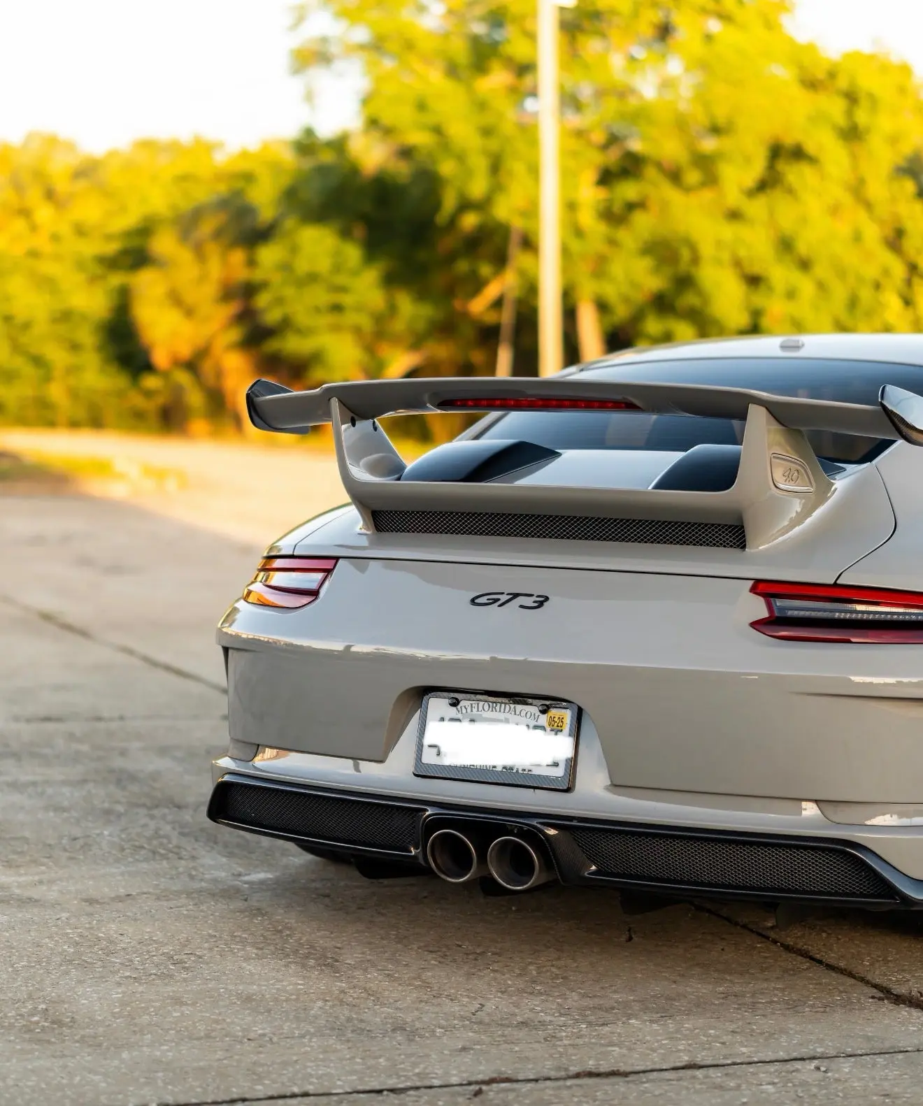 Rear view of sleek white Porsche GT3 sports car with carbon fiber diffuser, dual exhaust pipes, and large aerodynamic rear wing on suburban road.