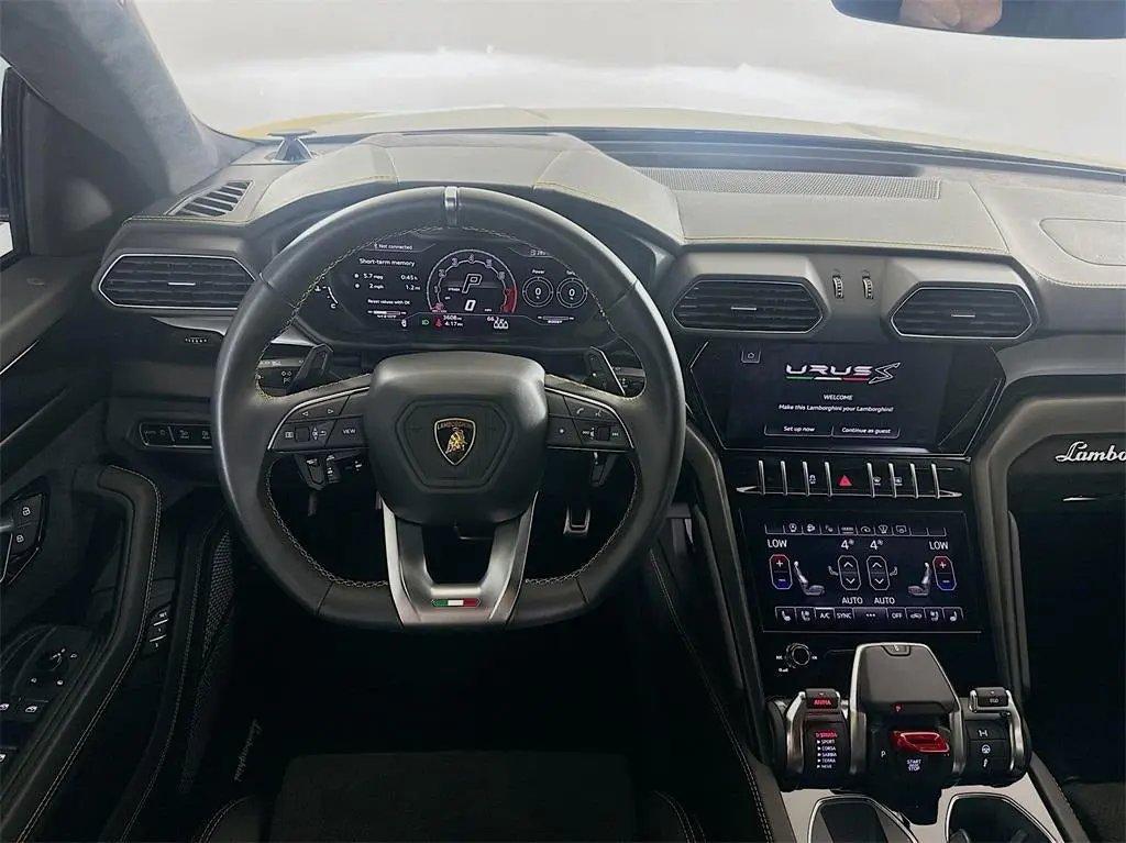 Interior view of Lamborghini Urus S luxury SUV showing black leather steering wheel with logo, digital dashboard display, and touchscreen infotainment system.