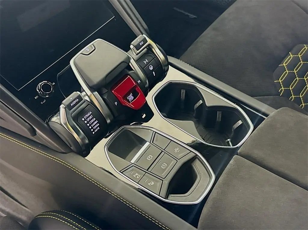 Close-up of luxury car center console with advanced gear shifter, driving mode selector, electronic parking brake, and dual cup holders, featuring black and yellow contrast stitching interior.