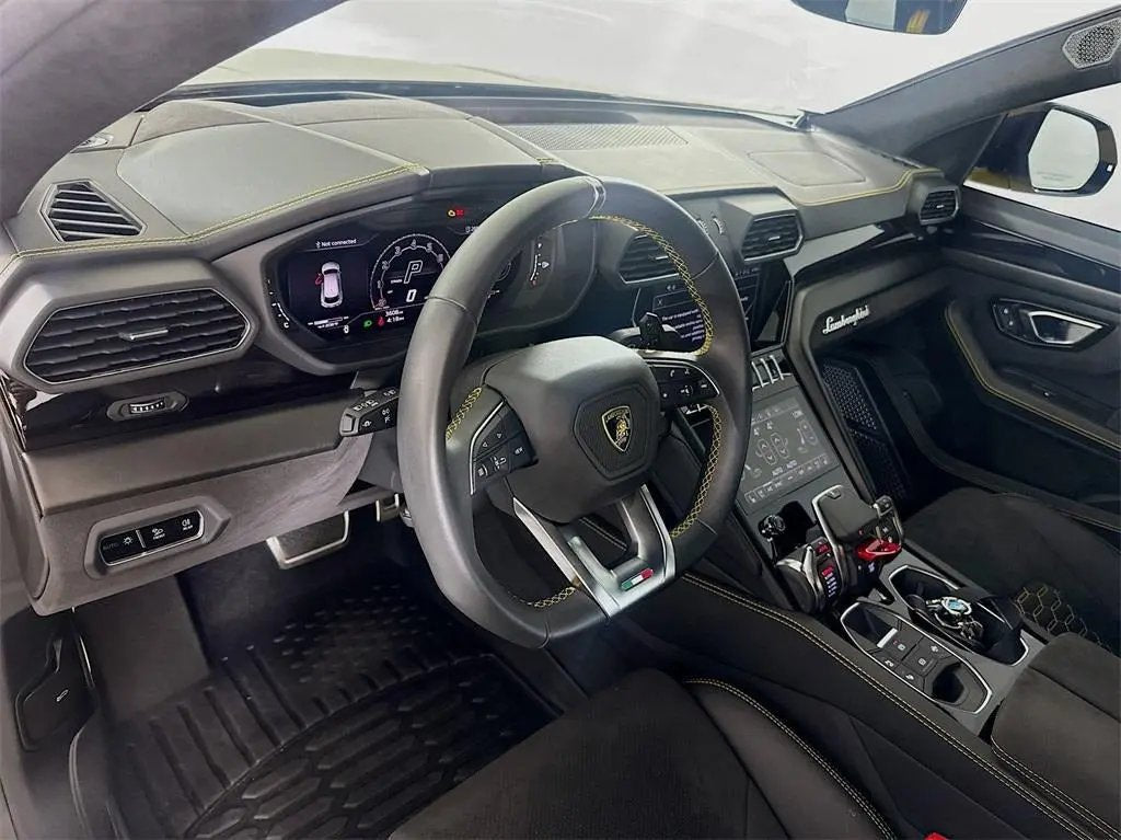 Luxury Lamborghini Urus interior with black leather steering wheel, digital dashboard, touchscreen controls, and premium black upholstery with yellow stitching.