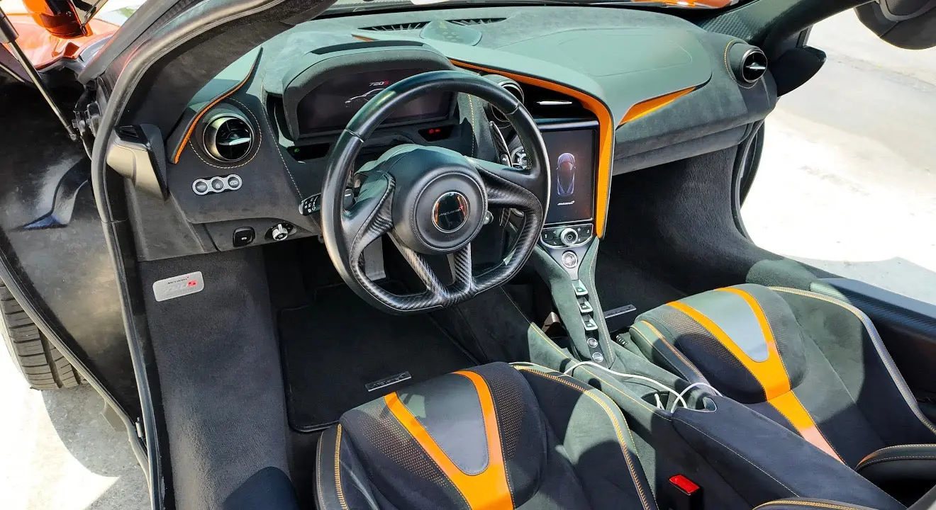 Interior view of McLaren 720S luxury sports car featuring black and orange leather seats, carbon fiber steering wheel, and high-tech dashboard with touchscreen display.