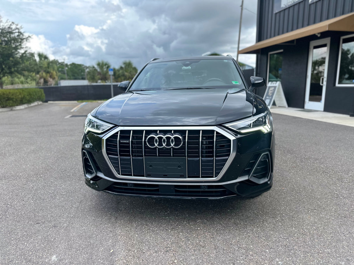 Alt text: Front view of black Audi Q3 luxury compact SUV with LED headlights and chrome grille, parked on asphalt near modern building.