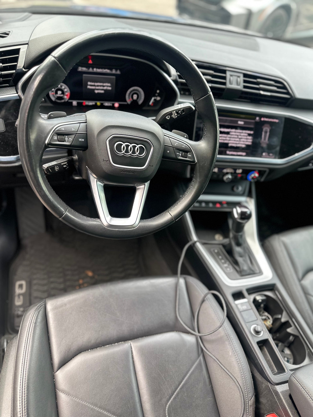 Alt text: Close-up of luxury Audi Q3 black leather steering wheel and driver seat interior with touchscreen display and gear shift in modern vehicle dashboard.