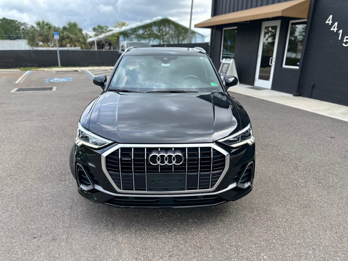 Black Audi Q3 SUV front view with LED headlights and chrome grille, parked outside showroom, luxury compact crossover car.