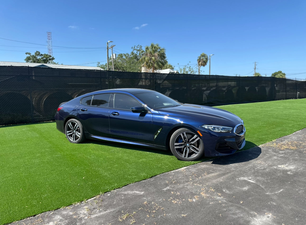 Sleek dark blue luxury BMW sedan parked on green artificial grass under clear blue sky, showcasing modern design and alloy wheels.