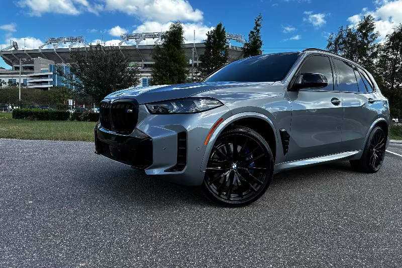 2023 BMW X7 M50i luxury SUV in metallic gray with black rims parked on asphalt under blue sky, high-performance sports SUV with sleek design.
