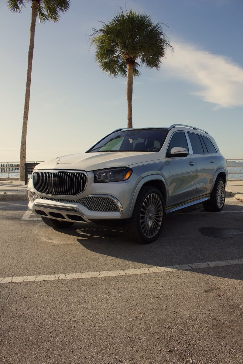 White luxury Mercedes-Benz GLS SUV with chrome grille and alloy wheels parked by palm trees near waterfront under blue sky