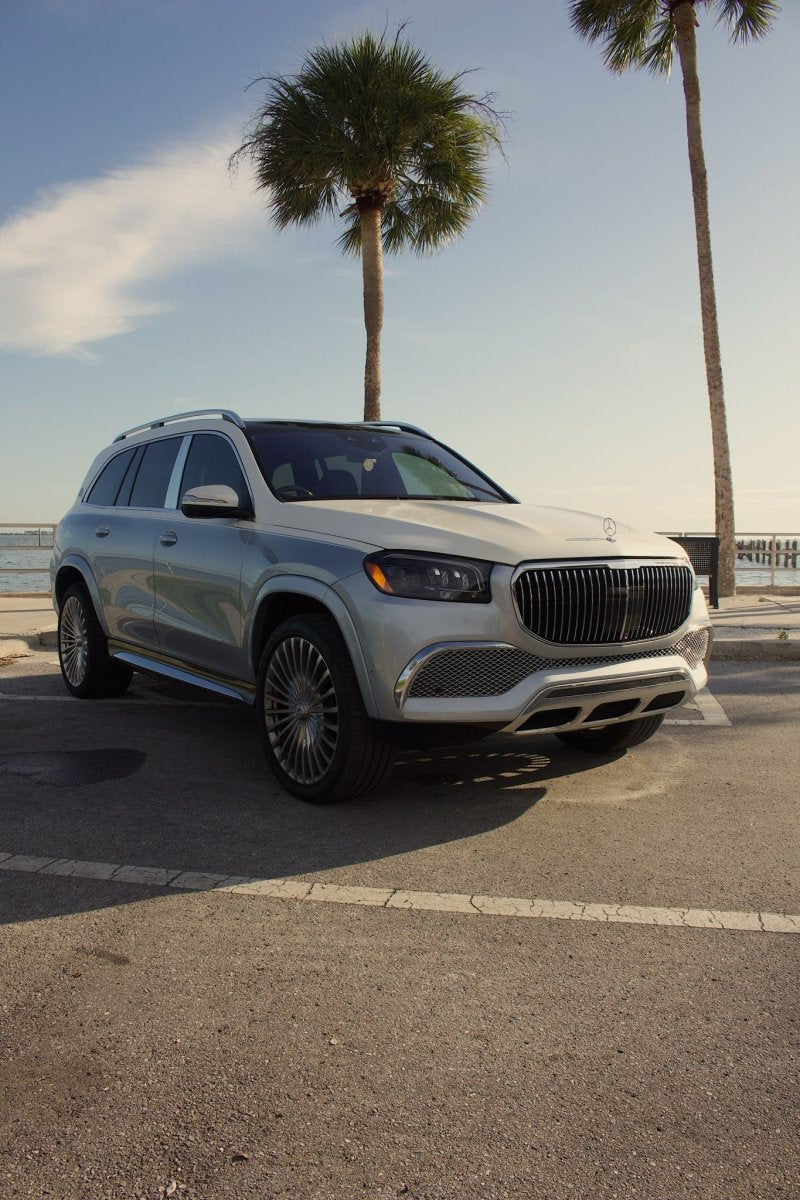 White luxury Mercedes-Benz GLS SUV with chrome grille and alloy wheels parked by palm trees near waterfront under blue sky